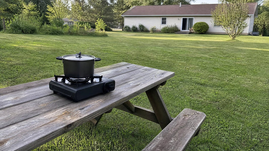 A small portable camp stove with a lidded pot on a wooden picnic table in an open grassy backyard, with a house across the lawn in the background