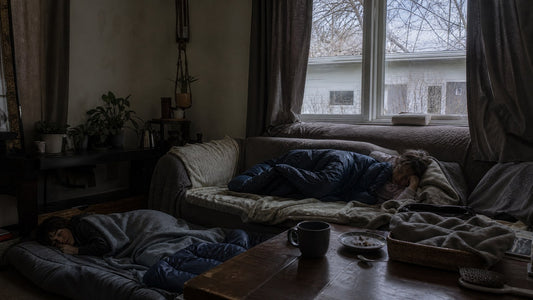 A living room on a gray winter afternoon with a couch piled with wool blankets and sleeping bags and curtains drawn